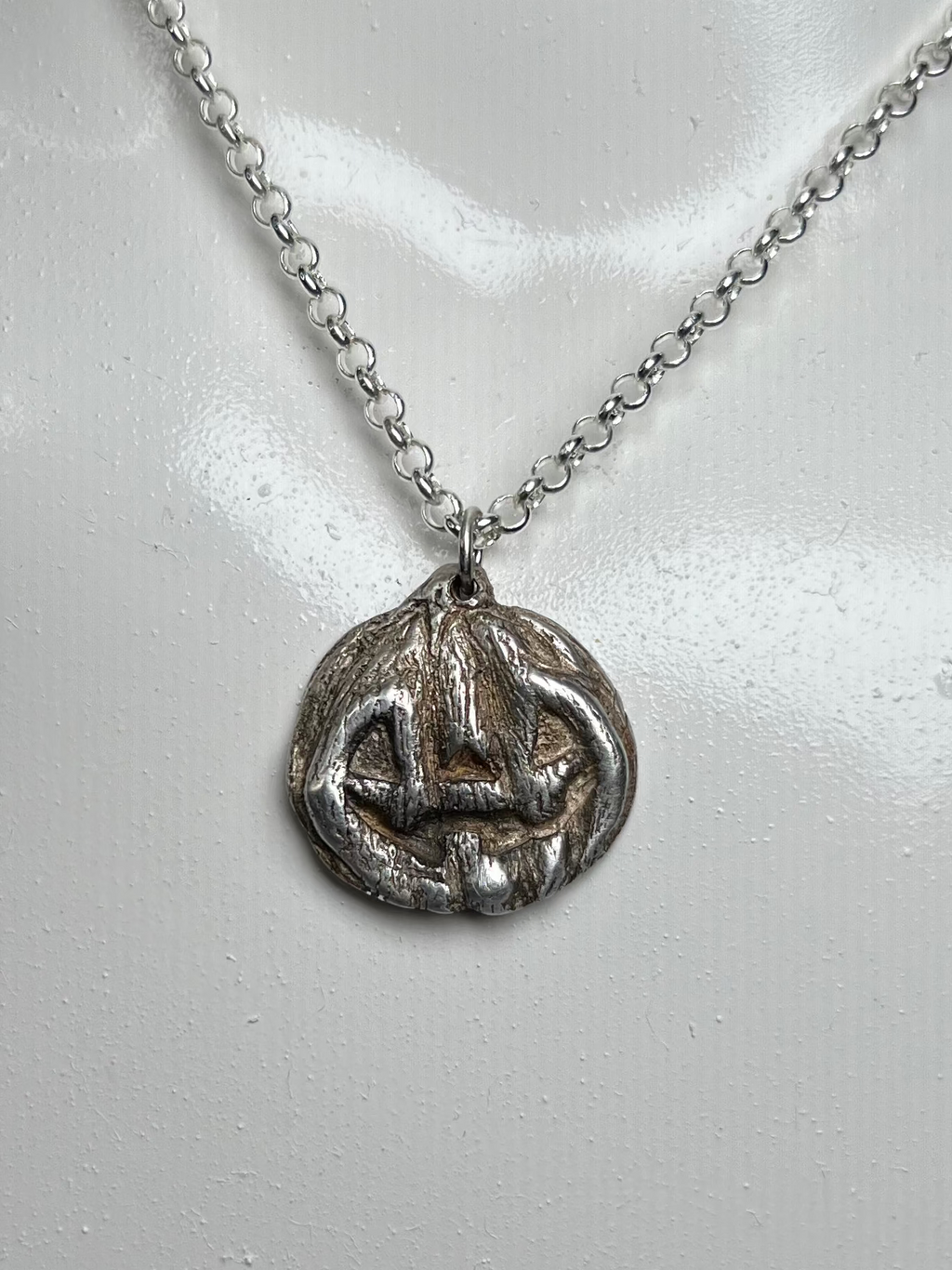 A silver pendant necklace displayed on a white mannequin bust against a dark background. The pendant appears to be a circular, textured jackolantern charm.