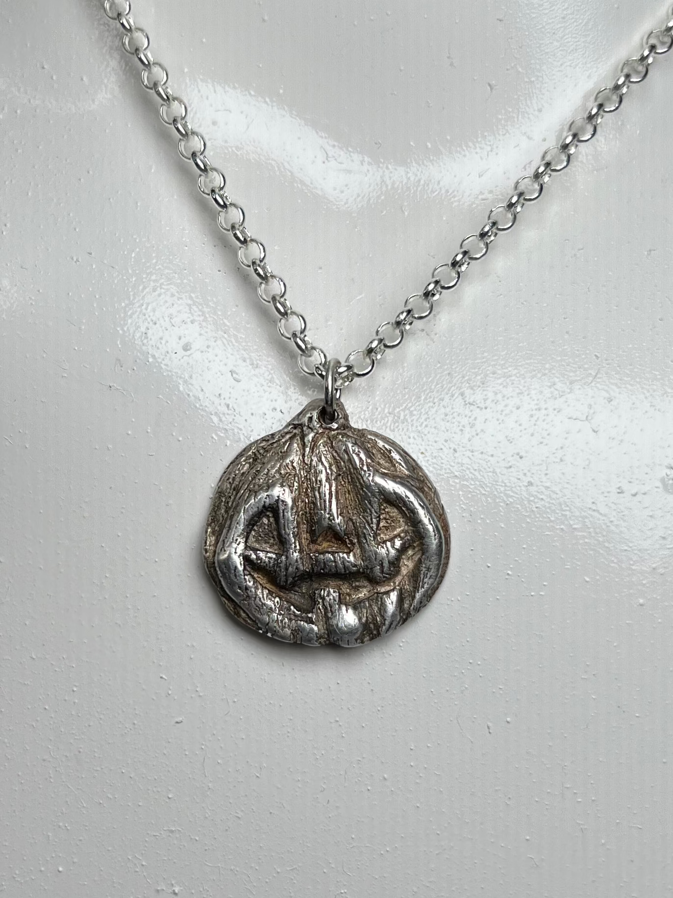 A silver pendant necklace displayed on a white mannequin bust against a dark background. The pendant appears to be a circular, textured jackolantern charm.