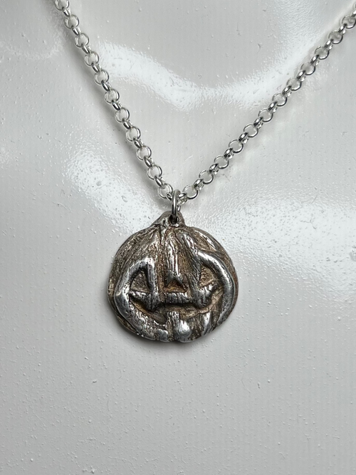 A silver pendant necklace displayed on a white mannequin bust against a dark background. The pendant appears to be a circular, textured jackolantern charm.